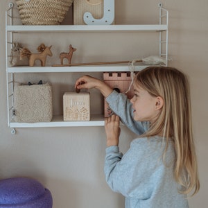 May include: A young girl is putting a coin into a wooden piggy bank with the name "Juna" on it. The piggy bank is on a white shelf with other toys, including a unicorn, a deer, and a wooden letter J.
