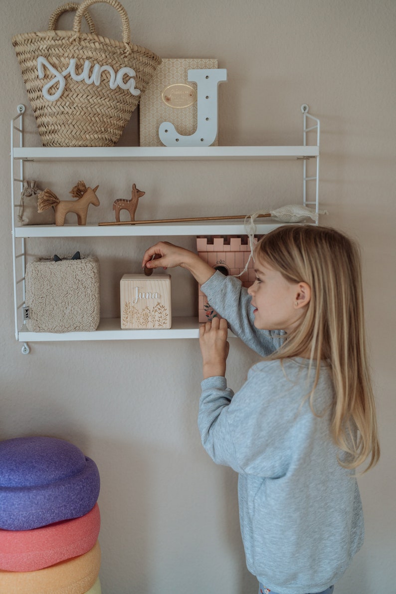 May include: A young girl is putting a coin into a wooden piggy bank with the name "Juna" on it. The piggy bank is on a white shelf with other toys, including a unicorn, a deer, and a wooden letter J.