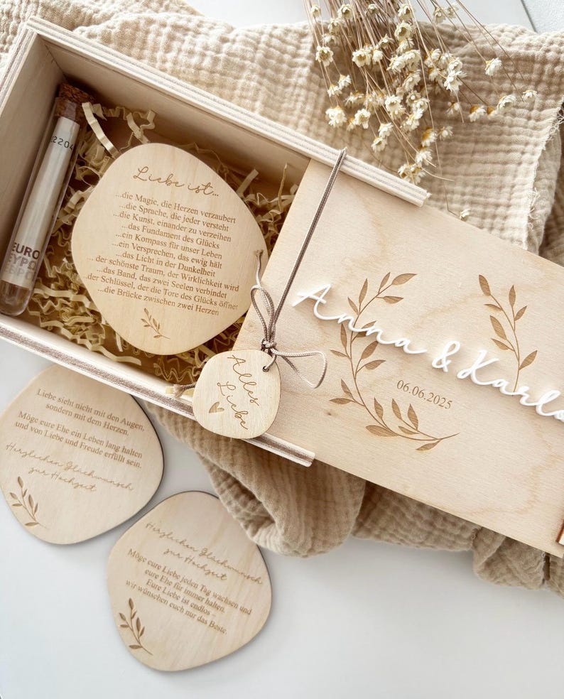 K&ouml;nnte beinhalten: Ein Holzbox-Set mit Gravurdetails, einschlie&szlig;lich eines Reagenzglases, herzf&ouml;rmigen Holzanh&auml;ngern und einem Deckel mit den Namen "Anna & Karla" und dem Datum "06.06.2025". Das Set zeigt florale Designs und deutschen Text.