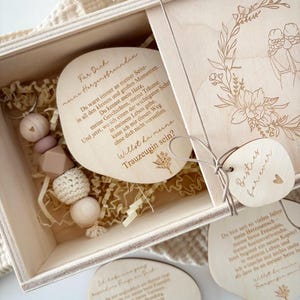 May include: A wooden gift box with a sliding lid, containing several wooden tags and a beaded accessory. The tags have engraved text and floral designs. The box is filled with shredded paper for cushioning. The overall theme is a personalized gift.