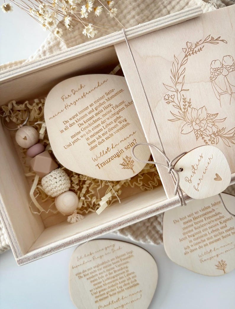 May include: A wooden gift box with a sliding lid, containing several wooden tags and a beaded accessory. The tags have engraved text and floral designs. The box is filled with shredded paper for cushioning. The overall theme is a personalized gift.