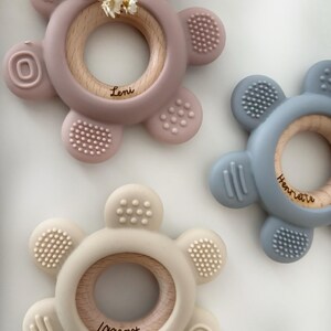 May include: Three silicone teethers with wooden rings in the center. The teethers are shaped like flowers with five petals. The teethers are in pink, blue, and beige. The wooden rings have the names "Leni", "Henriette", and "Lennart" engraved on them.