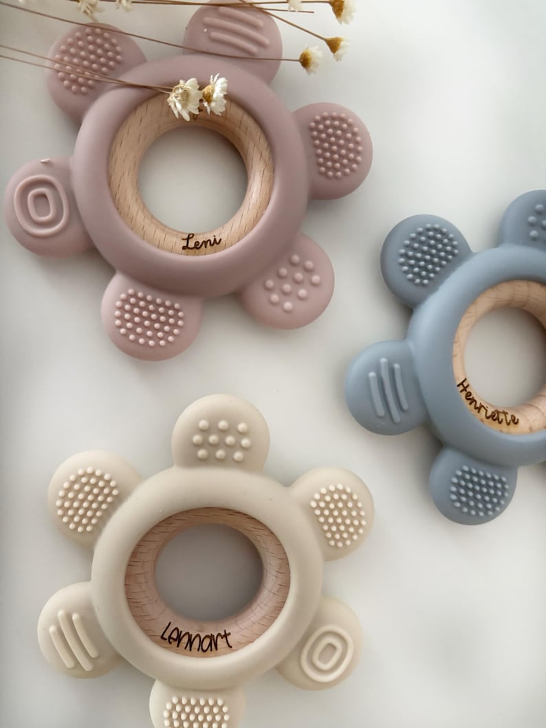 May include: Three silicone teethers with wooden rings in the center. The teethers are shaped like flowers with five petals. The teethers are in pink, blue, and beige. The wooden rings have the names "Leni", "Henriette", and "Lennart" engraved on them.