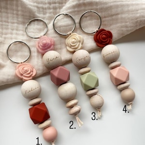 May include: Four personalized keychains with wooden beads, geometric shapes, and silicone flowers in shades of pink, red, and green. Each keychain has a different name engraved on it: Nadine, Yana, Sanja, and Bianca.