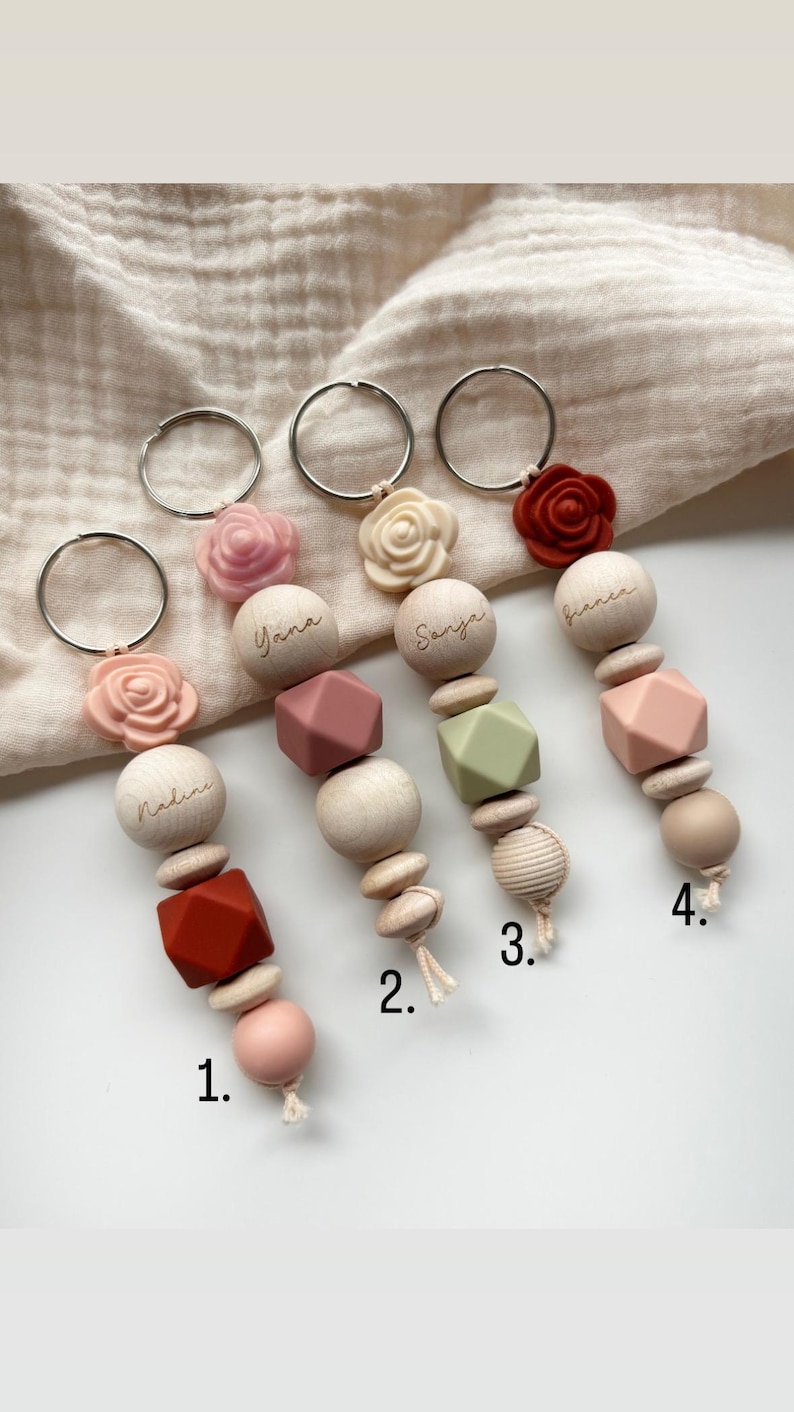 May include: Four personalized keychains with wooden beads, geometric shapes, and silicone flowers in shades of pink, red, and green. Each keychain has a different name engraved on it: Nadine, Yana, Sanja, and Bianca.
