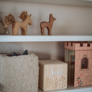 May include: A white wooden shelf with a wooden box that says "Juna" on it. The box is decorated with a floral design. There are also wooden animal figurines on the shelf, including a deer and a unicorn.