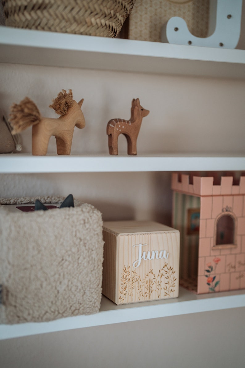 May include: A white wooden shelf with a wooden box that says "Juna" on it. The box is decorated with a floral design. There are also wooden animal figurines on the shelf, including a deer and a unicorn.