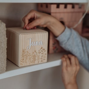 May include: A wooden box with the name "Juna" engraved on the front. The box has a floral design and is being placed on a white shelf.