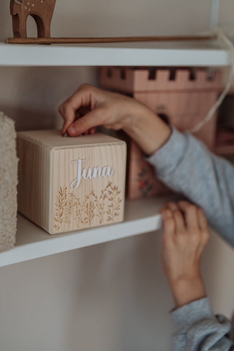 May include: A wooden box with the name "Juna" engraved on the front. The box has a floral design and is being placed on a white shelf.