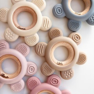 May include: Five silicone teethers in different colors with wooden rings in the center. The teethers have textured surfaces and are personalized with names: Emma, Philippe, Leni, Alma, and Lili.