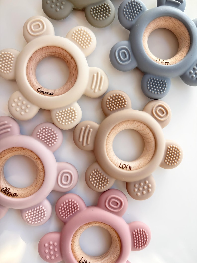 May include: Five silicone teethers in different colors with wooden rings in the center. The teethers have textured surfaces and are personalized with names: Emma, Philippe, Leni, Alma, and Lili.