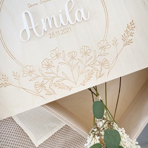 Puede incluir: Una caja de madera para recuerdos con un recorte circular y un dise&ntilde;o floral. La caja est&aacute; grabada con el texto "Deine Erinnerungen" y el nombre "Amila" con la fecha "28.11.2021".
