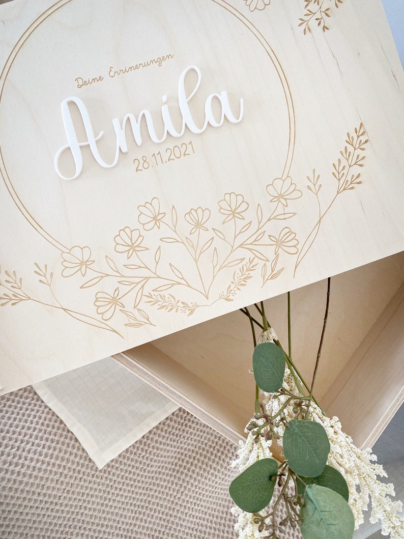 Puede incluir: Una caja de madera para recuerdos con un recorte circular y un dise&ntilde;o floral. La caja est&aacute; grabada con el texto "Deine Erinnerungen" y el nombre "Amila" con la fecha "28.11.2021".