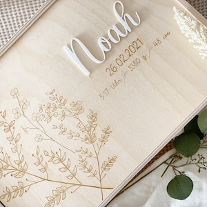 Könnte beinhalten: Eine Holzkiste mit einem floralen Design, das in den Deckel graviert ist. Der Deckel trägt außerdem den Namen "Noah" und das Datum "26.02.2021" sowie die Uhrzeit "5:17 Uhr", das Gewicht "3380 g" und die Länge "48 cm", die in den Deckel graviert sind.