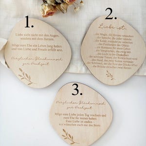 K&ouml;nnte beinhalten: Drei Hochzeitsgeschenke aus Holz mit abgerundeten Kanten, jeweils mit deutschem Text und botanischen Illustrationen graviert. Die Geschenke zeigen verschiedene romantische Zitate und Gl&uuml;ckw&uuml;nsche f&uuml;r das Paar. Trockenblumen sind im Hintergrund.