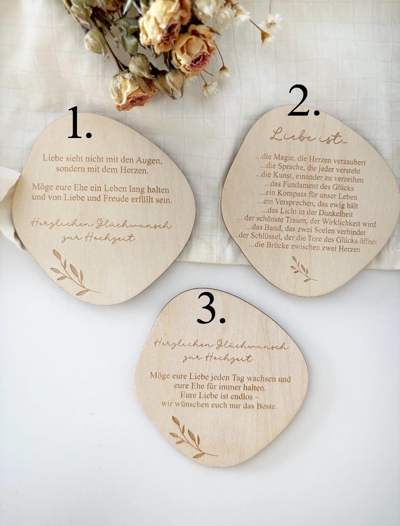 K&ouml;nnte beinhalten: Drei Hochzeitsgeschenke aus Holz mit abgerundeten Kanten, jeweils mit deutschem Text und botanischen Illustrationen graviert. Die Geschenke zeigen verschiedene romantische Zitate und Gl&uuml;ckw&uuml;nsche f&uuml;r das Paar. Trockenblumen sind im Hintergrund.
