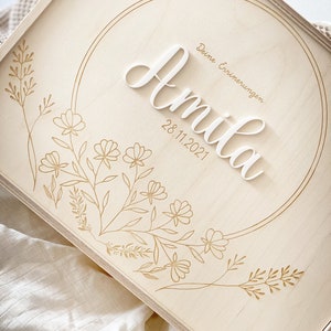 Puede incluir: Una caja de madera clara con un dise&ntilde;o circular grabado en la parte delantera. El c&iacute;rculo contiene el nombre "Amilia" en letras blancas y la fecha "28.11.2021" debajo. El texto "Deine Erinnerungen" est&aacute; escrito encima del nombre. La caja est&aacute; decorada con un dise&ntilde;o floral.