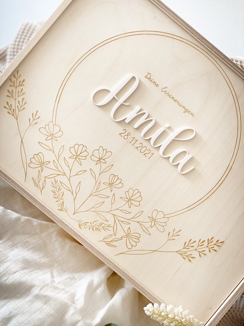 Puede incluir: Una caja de madera clara con un dise&ntilde;o circular grabado en la parte delantera. El c&iacute;rculo contiene el nombre "Amilia" en letras blancas y la fecha "28.11.2021" debajo. El texto "Deine Erinnerungen" est&aacute; escrito encima del nombre. La caja est&aacute; decorada con un dise&ntilde;o floral.
