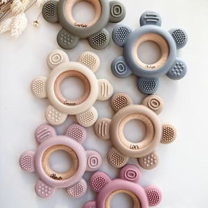 May include: Five silicone teethers in different colors with wooden rings in the center. Each teether has a different name engraved on the ring: Carlos, Philippa, Emma,  Levi, and Lily.