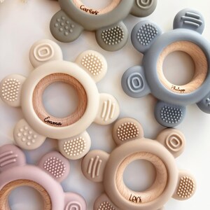 May include: Five silicone teethers with wooden rings in the center. The teethers are in different colors: green, blue, beige, pink, and light brown. Each teether has a different name engraved on the wooden ring: Carlos, Philip, Emma, Leni, and Alma.