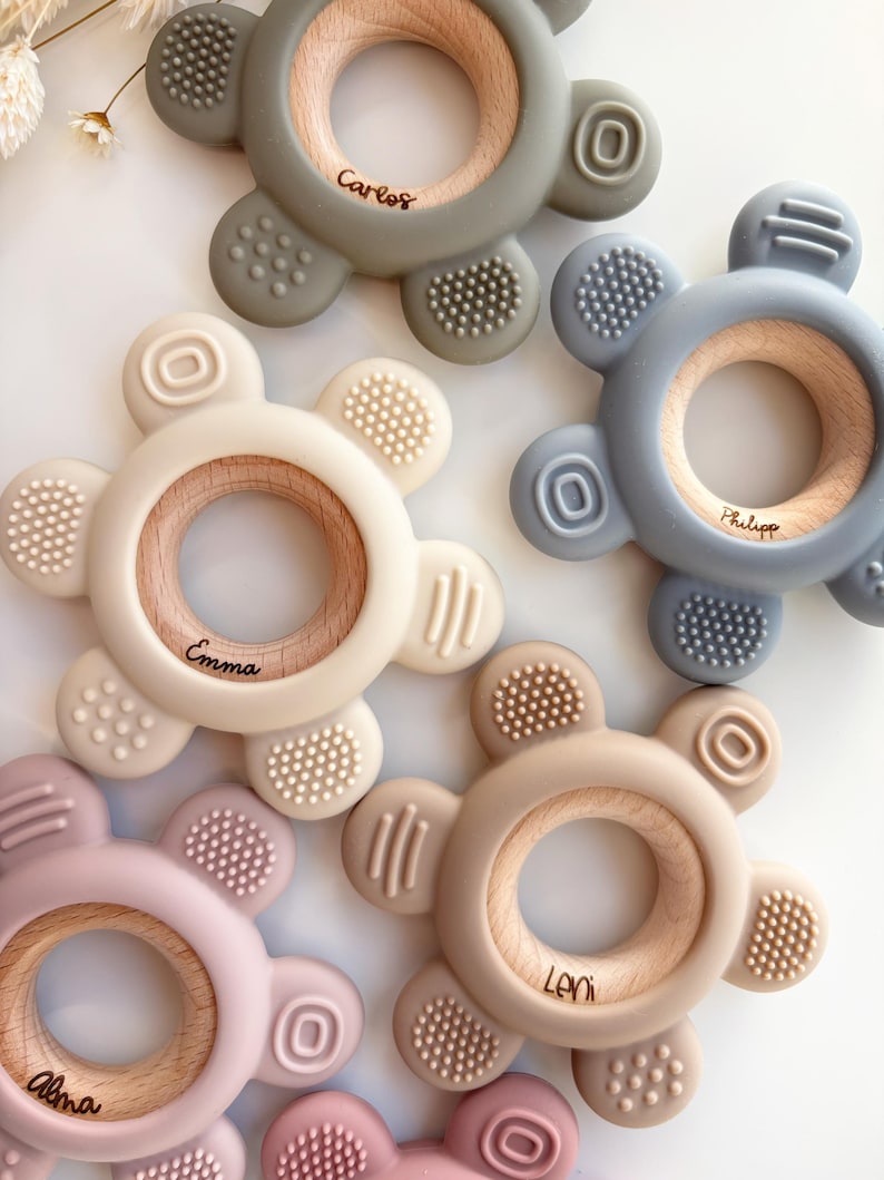 May include: Five silicone teethers with wooden rings in the center. The teethers are in different colors: green, blue, beige, pink, and light brown. Each teether has a different name engraved on the wooden ring: Carlos, Philip, Emma, Leni, and Alma.