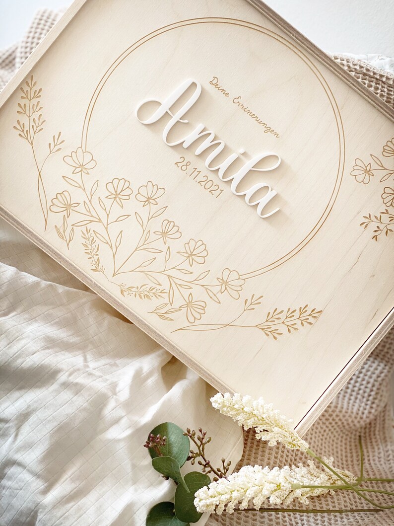 Puede incluir: Una caja de madera con fondo blanco y dise&ntilde;os florales dorados. La caja tiene un c&iacute;rculo en el centro con el nombre "Amilia" y la fecha "28.11.2021" escritos en blanco. El texto "Deine Erinnerungen" est&aacute; escrito en dorado sobre el nombre.