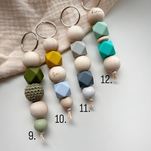 May include: Four keychains with wooden beads and colorful silicone beads. Each keychain has a silver ring. The beads are in various shapes and colors, including green, yellow, gray, and blue. Some keychains have a name engraved on a wooden bead.