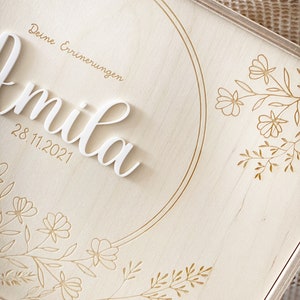 Puede incluir: Una caja de madera con una inscripci&oacute;n blanca que dice "Deine Erinnerungen" y "mila" con la fecha "28.11.2021". La caja est&aacute; decorada con un c&iacute;rculo dorado y dise&ntilde;os florales.