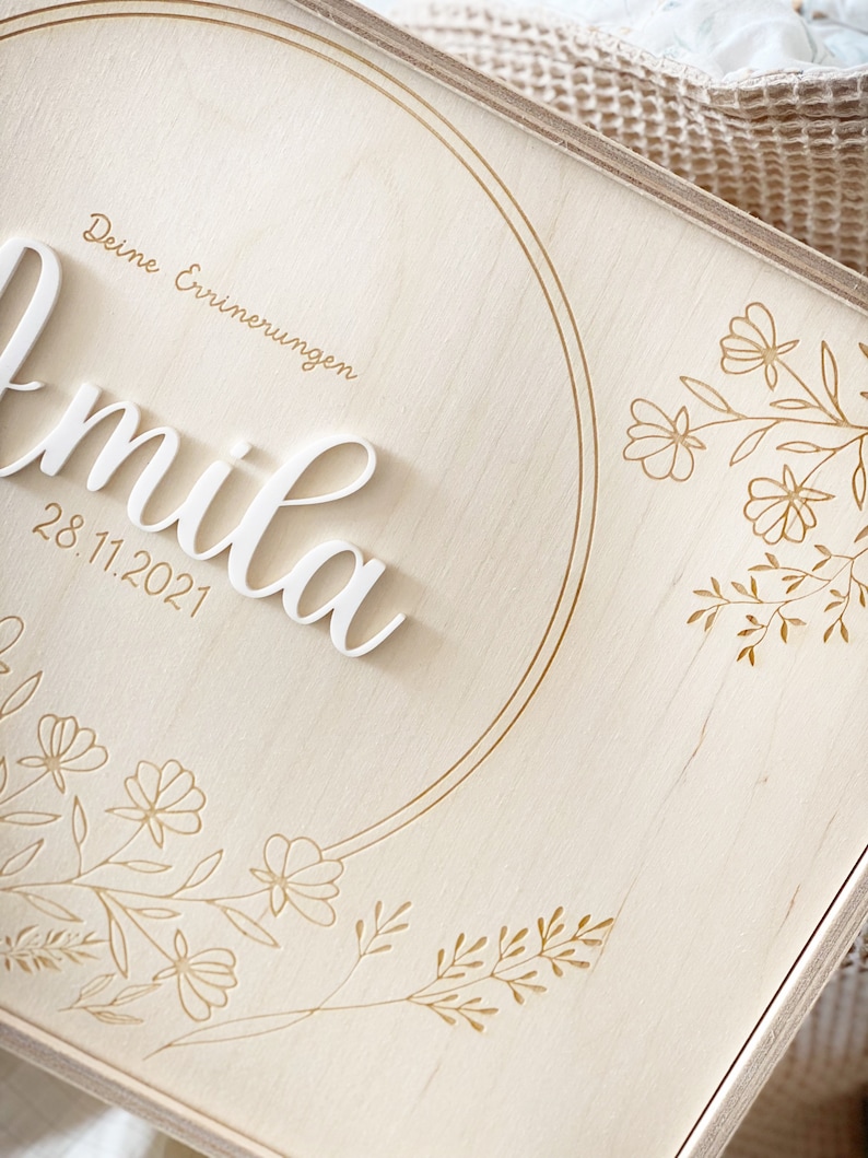 Puede incluir: Una caja de madera con una inscripci&oacute;n blanca que dice "Deine Erinnerungen" y "mila" con la fecha "28.11.2021". La caja est&aacute; decorada con un c&iacute;rculo dorado y dise&ntilde;os florales.