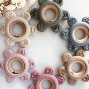 May include: Six silicone teethers in different colors with wooden rings in the center. Each teether has a different name engraved on the ring: Carlos, Emma, Alina, Liam, Lily, and Philip.