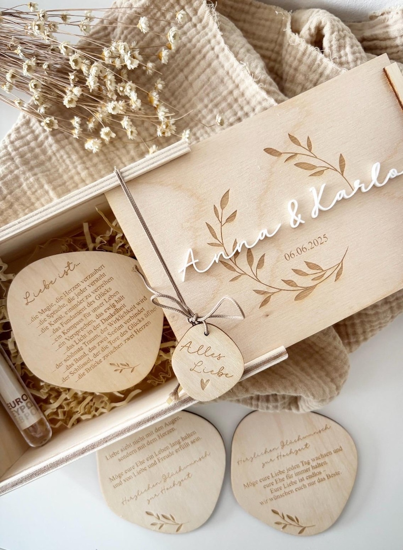 K&ouml;nnte beinhalten: Eine Holzgeschenkbox mit den Namen "Anna & Karla" und dem Datum "06.06.2025" auf dem Deckel. Im Inneren befinden sich Holzuntersetzer und ein kleines Fl&auml;schchen. Ein Holzanh&auml;nger mit "Alles Liebe" ist befestigt.