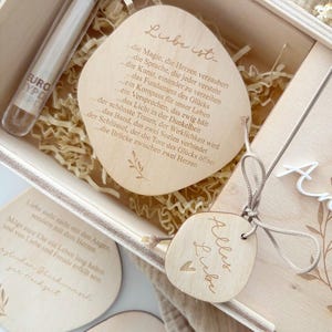 K&ouml;nnte beinhalten: Holzgeschenkbox mit einem transparenten Glasr&ouml;hrchen und mehreren Holzanh&auml;ngern. Die Anh&auml;nger haben gravierten Text und ein Herzdesign. Die Box enth&auml;lt Dekopapier und einen Holzdeckel mit floralen Akzenten.