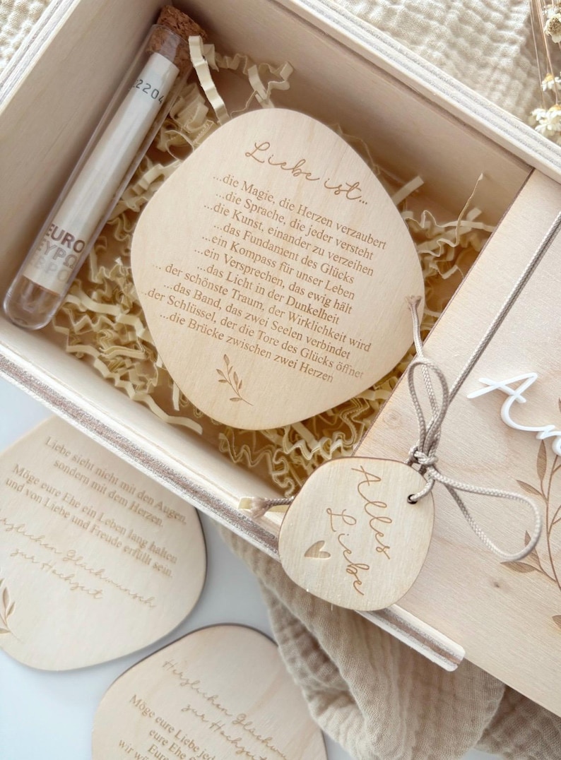 K&ouml;nnte beinhalten: Holzgeschenkbox mit einem transparenten Glasr&ouml;hrchen und mehreren Holzanh&auml;ngern. Die Anh&auml;nger haben gravierten Text und ein Herzdesign. Die Box enth&auml;lt Dekopapier und einen Holzdeckel mit floralen Akzenten.