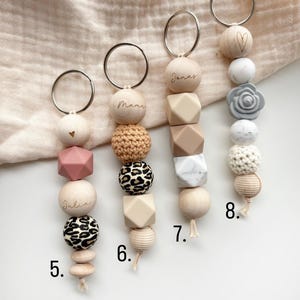 May include: A collection of baby pacifier clips with various designs. Each clip features a metal ring, wooden beads, and silicone beads in different shapes and colors. Some clips have names like "Julia" and "Janas" engraved on them, while others have decorative elements like a heart or a flower.