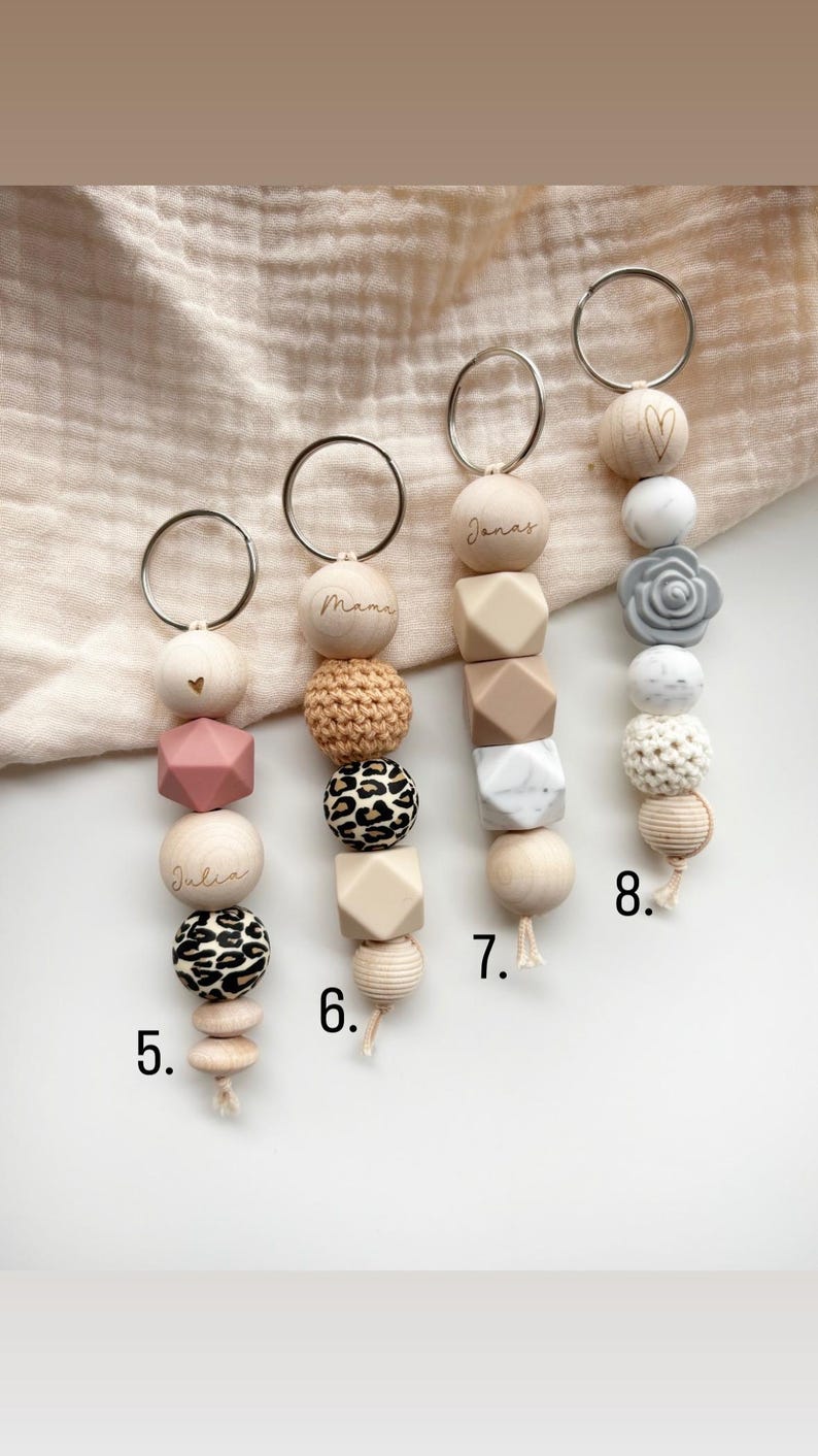 May include: A collection of baby pacifier clips with various designs. Each clip features a metal ring, wooden beads, and silicone beads in different shapes and colors. Some clips have names like "Julia" and "Janas" engraved on them, while others have decorative elements like a heart or a flower.