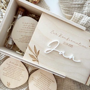 May include: A wooden box with a sliding lid, featuring the inscription "Zur Konfirmation" and the name "Jan" with the date 20.04.2025. Inside are two small glass vials and two wooden coasters with printed text and leaf designs. The box is made of light-colored wood.
