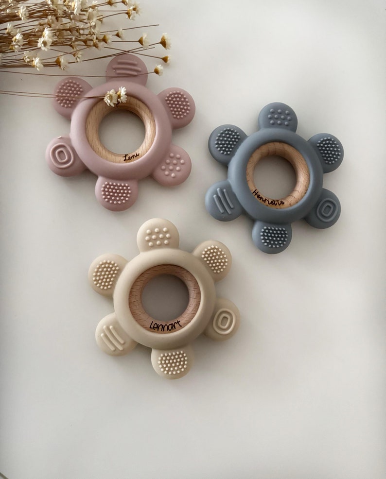 May include: Three silicone teethers in pink, gray, and beige with a wooden ring in the center. Each teether has a different name engraved on the ring: "Leni", "Henriette", and "Lennart".