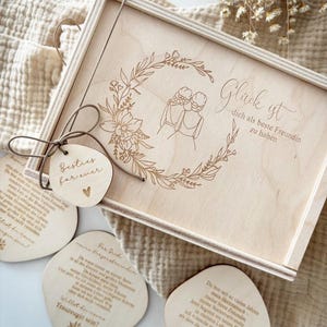 May include: A wooden gift box with a floral wreath design and the words "Gl&uuml;ck ist dich als beste Freundin zu haben" engraved on the lid. Includes wooden tags with text and a small heart. The box is tied with a brown string.