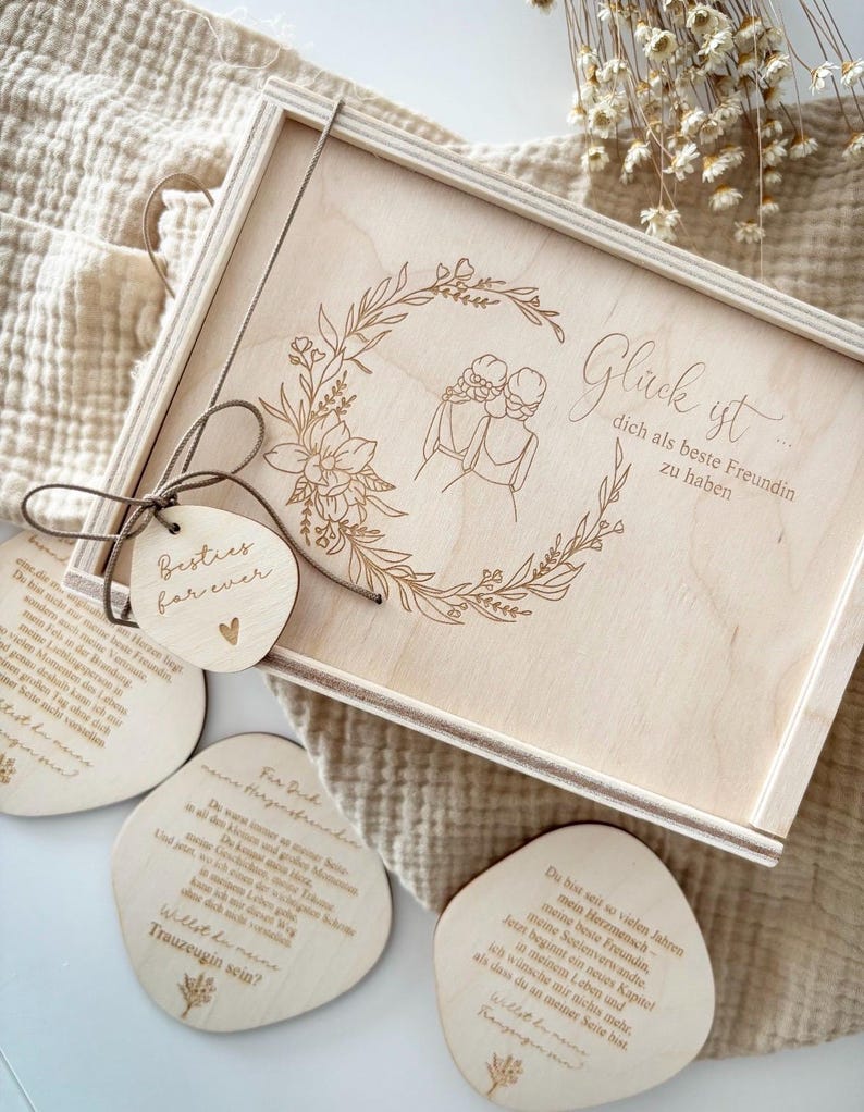 May include: A wooden gift box with a floral wreath design and the words "Gl&uuml;ck ist dich als beste Freundin zu haben" engraved on the lid. Includes wooden tags with text and a small heart. The box is tied with a brown string.