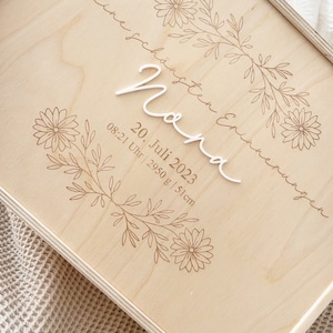 Könnte beinhalten: Eine Holzkiste mit floralem Design und dem Text "Meine schönste Erinnerung" und "Nora" in weißer Schrift. Das Datum "20. Juli 2023" und die Uhrzeit "08:21 Uhr | 2950 g | 51 cm" sind ebenfalls in die Kiste graviert.