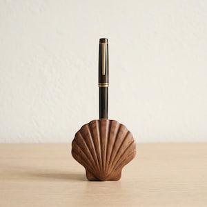 May include: A dark brown wooden pen holder carved into the shape of a scallop shell, featuring a vertical slot at the top center. A black fountain pen with a gold-colored clip and trim is inserted upright into the holder, resting securely in place.