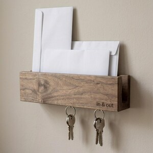 May include: A rectangular wooden wall-mounted mail organizer with a natural wood grain finish. The front panel features the text "in & out" engraved in the bottom right corner. Two metal rings hang from the bottom, each holding a set of silver keys.