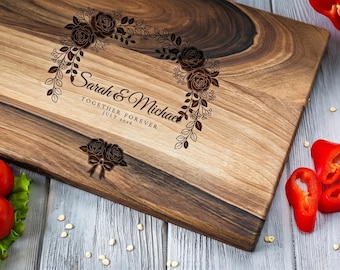 May include: A rectangular wooden cutting board with a dark and light wood grain pattern. It features a laser-engraved floral wreath design surrounding the text Sarah & Michael, Together Forever, July 2026, and a small rose bow graphic.