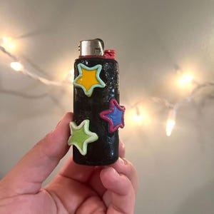 May include: Black lighter decorated with colorful star-shaped embellishments. The stars are outlined in white and filled with yellow, blue, green, and pink. The lighter has a silver metal top and a red ignition button.