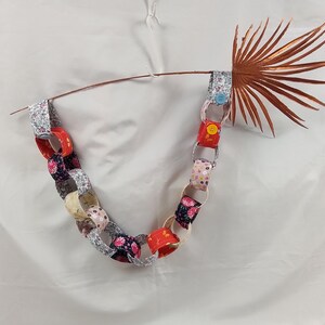 Könnte beinhalten: Eine bunte Papiergirlande mit gemusterten Gliedern in Rot, Rosa, Blumenmuster und Silber. Die Girlande ist über einem braunen Palmwedel drapiert und mit blauen und gelben Knöpfen befestigt. Der Hintergrund ist weiß.
