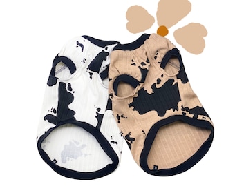 May include: Two dog shirts with a cow print pattern. One shirt is white with black spots and the other is brown with black spots.