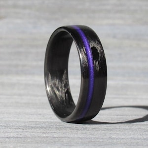 May include: A black ring with a purple stripe. The ring is made of a dark material and has a smooth, polished finish. The purple stripe runs through the centre of the ring.