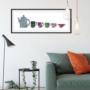 May include: A framed print of a blue teapot and five colourful mugs with floral and geometric patterns. The print is hanging on a white wall above a green sofa with orange cushions.