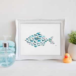 K&ouml;nnte beinhalten: Ein gerahmter Druck mit einem Fisch, der aus verschiedenen Illustrationen von blauen und t&uuml;rkisfarbenen Fischen besteht. Das Bild ist vor einem wei&szlig;en Hintergrund, mit einem Stapel t&uuml;rkisfarbener Handt&uuml;cher, einem Seifenspender, einer kleinen Pflanze und einer Gummiente.