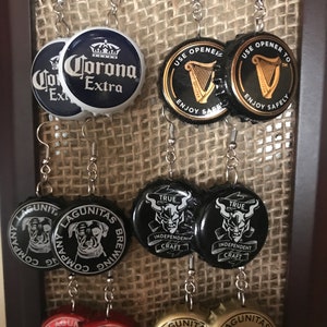 May include: Eight pairs of earrings made from bottle caps. The bottle caps are decorated with various logos and designs, including Corona Extra, Guinness, and Lagunitas Brewing Company. The earrings are silver and have a simple design.